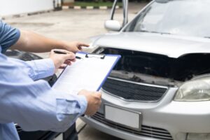 Insurance officer writing on a clipboard while an insurance agent examines a black car after an accident.