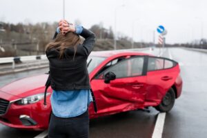 A woman stands near a broken car after an accident, calling for help and considering car insurance options.