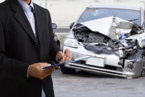 Side view of an insurance officer writing on a clipboard while an insurance agent examines a silver car after an accident.