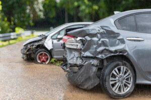 On a rainy day, a light gray car crashed into the back of a gray car, leaving both vehicles damaged and undriveable, parked on the road.