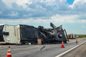 A fifth-wheel recreational vehicle overturned on a highway, with its underside torn apart and belongings scattered across the roadway after an accident.