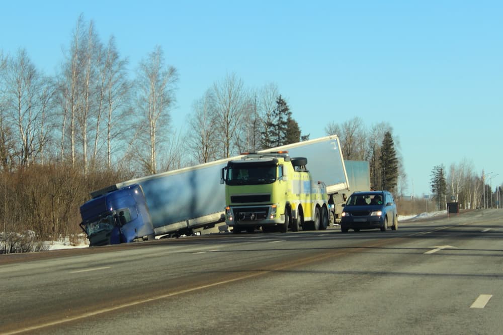 A heavily wrecked truck lies in a roadside ditch on a spring day, with an average commissioner on-site assessing the road transport accident.