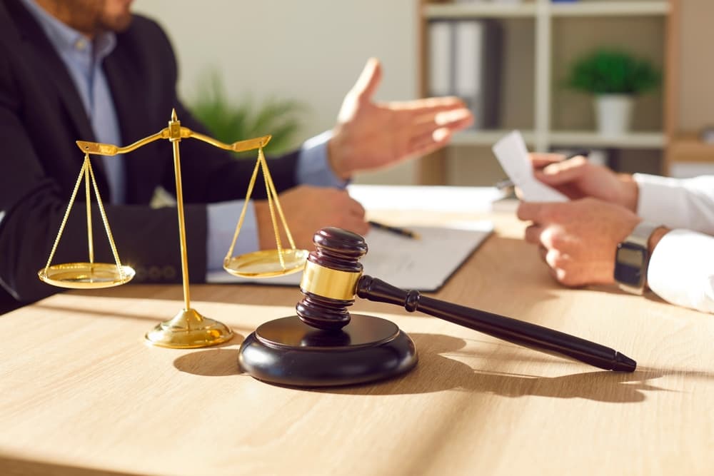 A close-up photo of a male lawyer working in his office, discussing with a male client at the desk, featuring a judge's gavel and scales in the background.